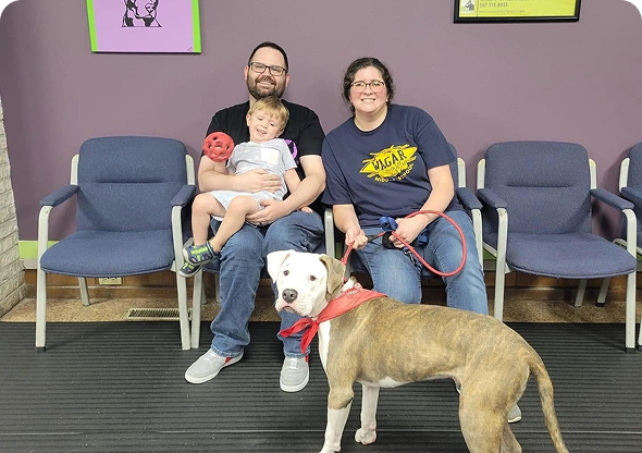 A family of three, including a young child, sits on chairs at a veterinary clinic. They have a large dog wearing a red bandana on a red leash. Purple walls and framed pictures are visible. A family of three, including a young child, sits on chairs at a veterinary clinic. They have a large dog wearing a red bandana on a red leash. Purple walls and framed pictures are visible.