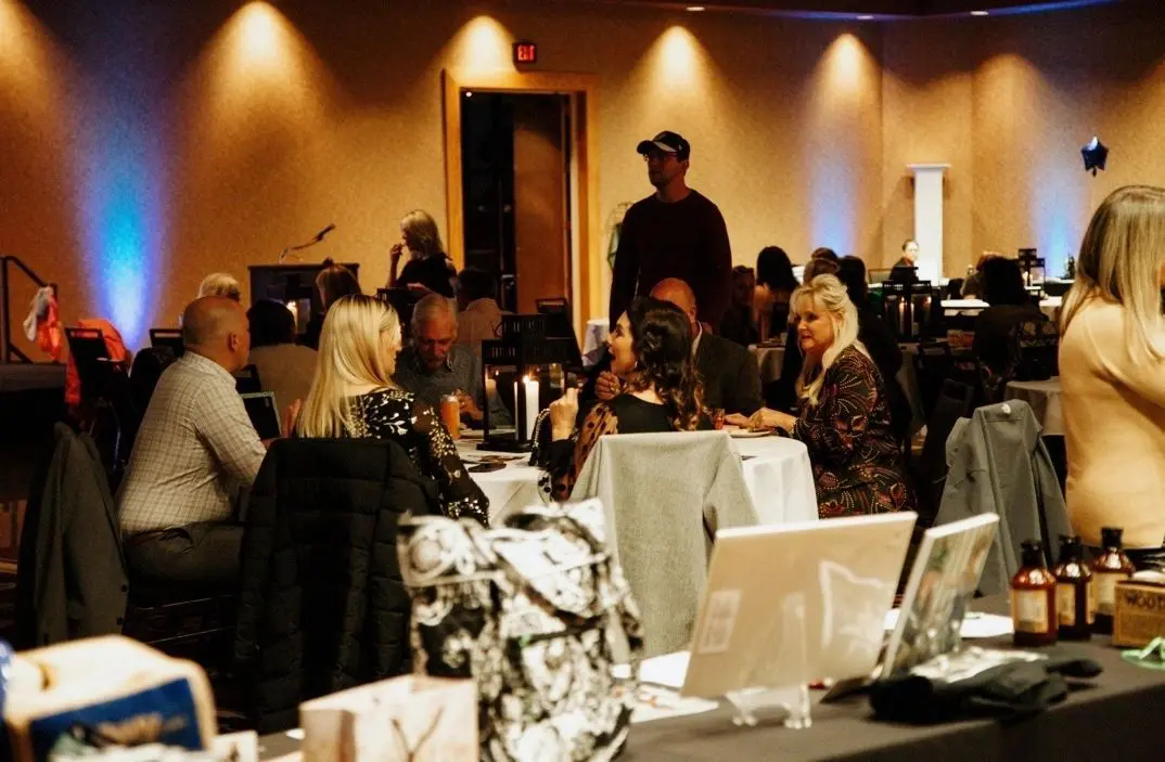 People sit at tables in a dimly lit room with decorated walls, engaging in conversation. A person in a cap stands in the background, and various items are displayed in the foreground.