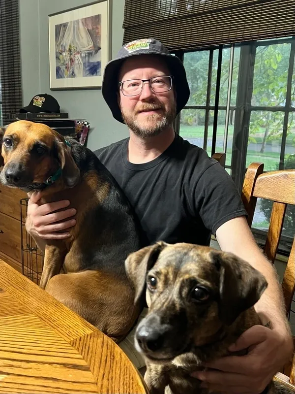 A man wearing a bucket hat sits at a wooden table indoors, holding two dogs on his lap. A window with blinds is in the background. A man wearing a bucket hat sits at a wooden table indoors, holding two dogs on his lap. A window with blinds is in the background.