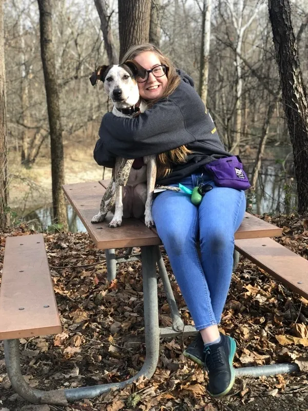 Person sitting on a picnic table in a forest, hugging a black and white dog. The table is covered with fallen leaves. The background shows trees and a river. Person sitting on a picnic table in a forest, hugging a black and white dog. The table is covered with fallen leaves. The background shows trees and a river.